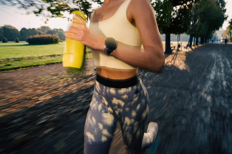 A woman running outside holding a yellow waterdrop® water bottle.