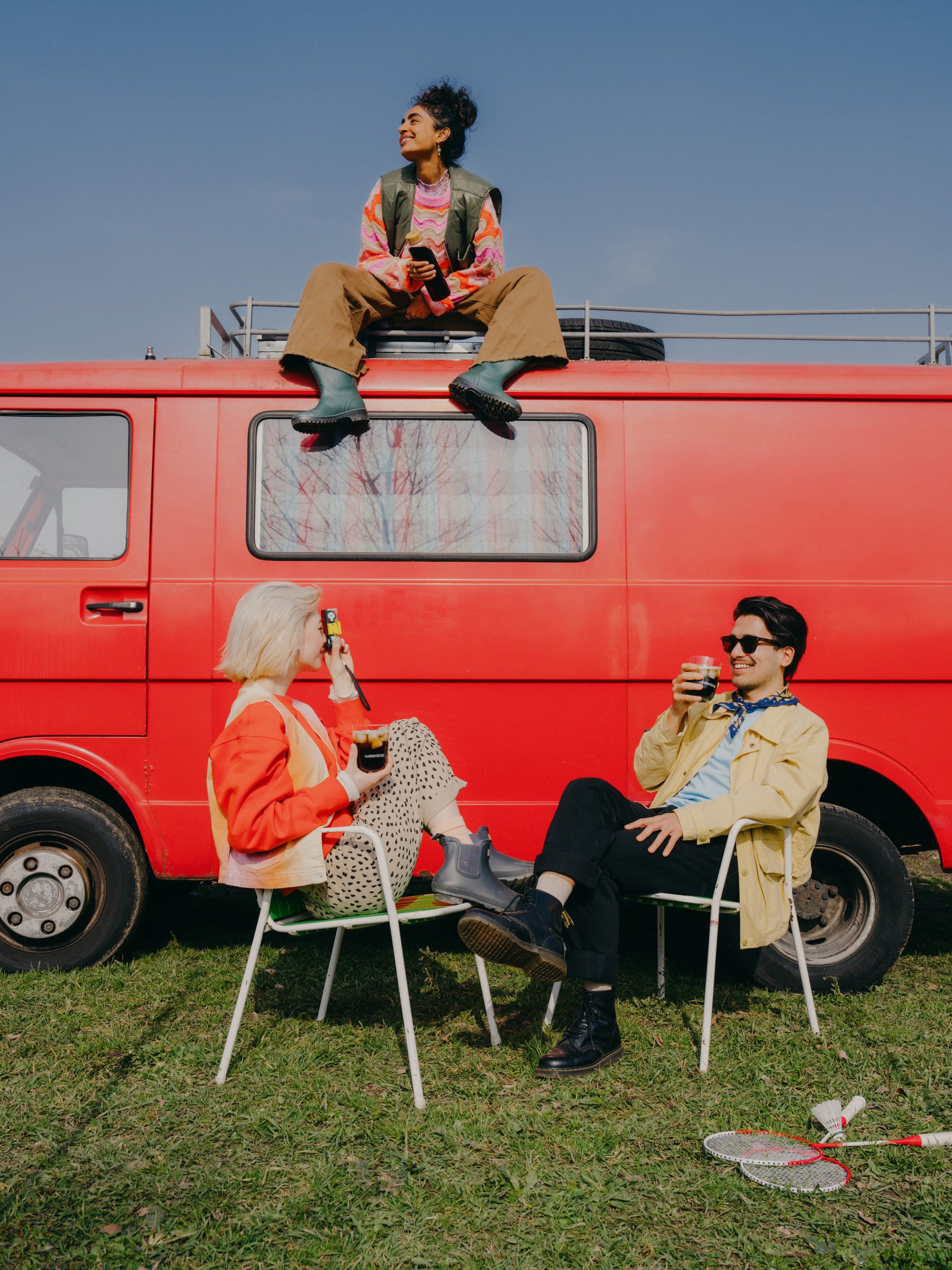A group of people sitting outside by a red van, enjoying drinks, with a waterdrop® branded water bottle visible.