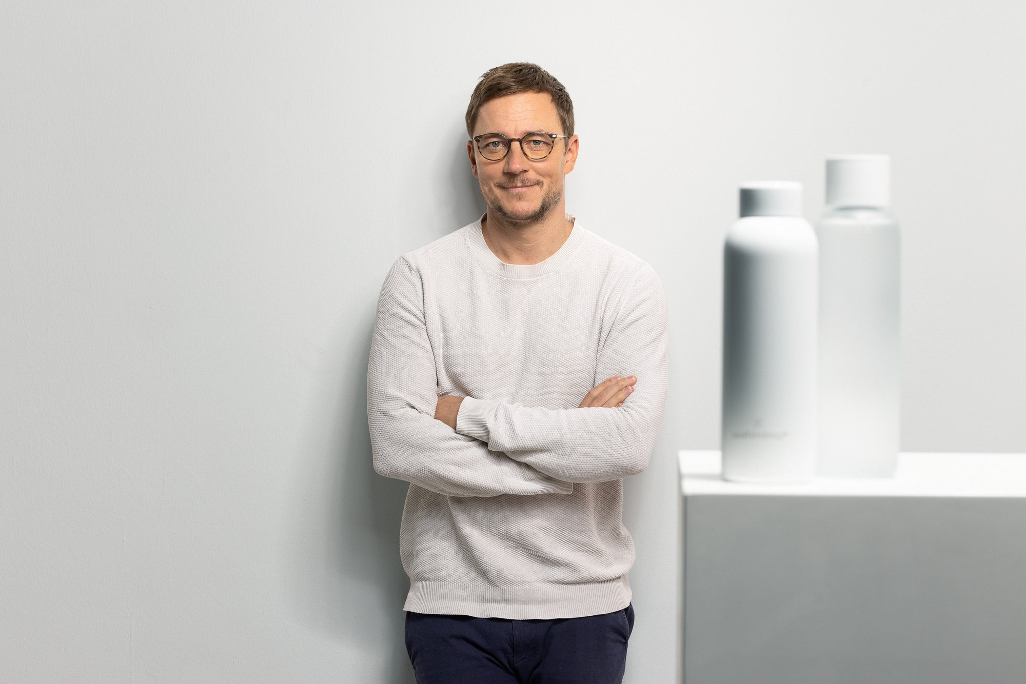 A man with glasses and a white shirt stands with his arms crossed, smiling.
