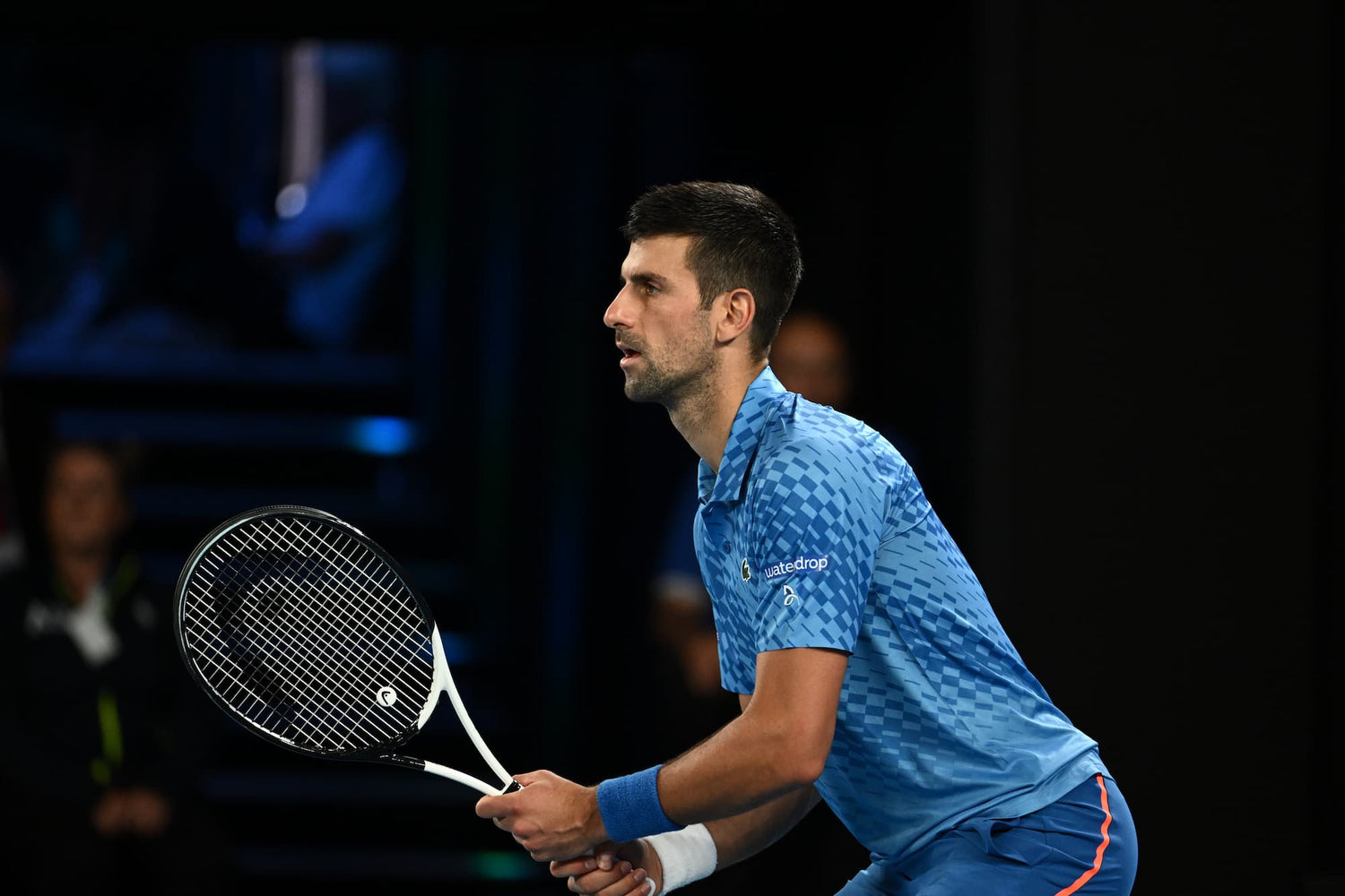 A man holding a tennis racket, wearing a blue shirt and blue wrist band, with short hair and a beard.