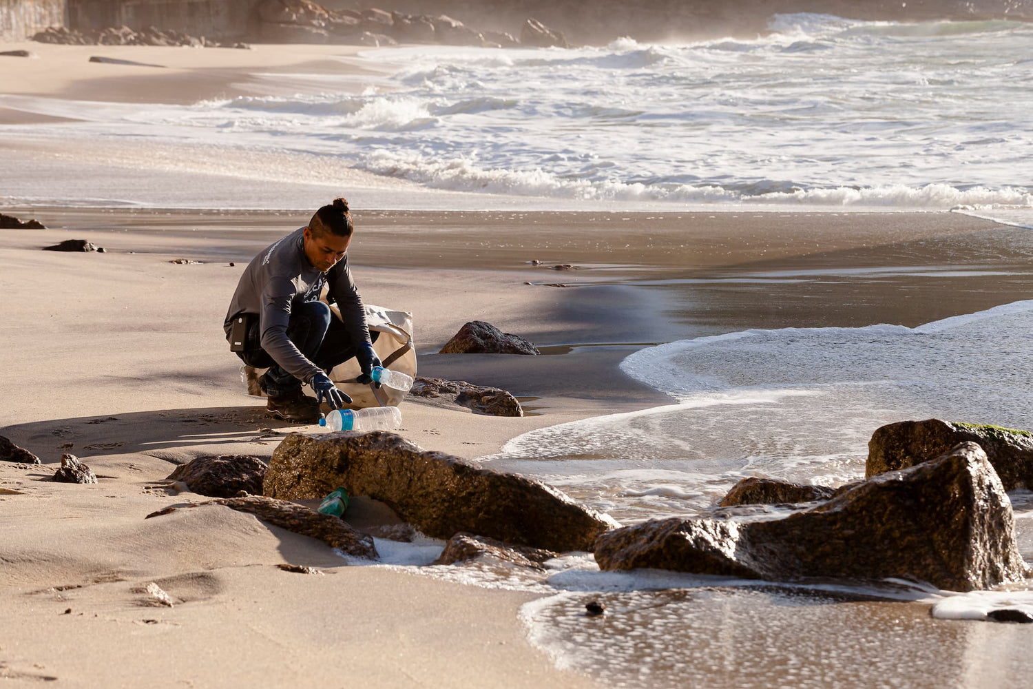 A man with a bun picking up plastic bottles on a beach with waves in the background.
