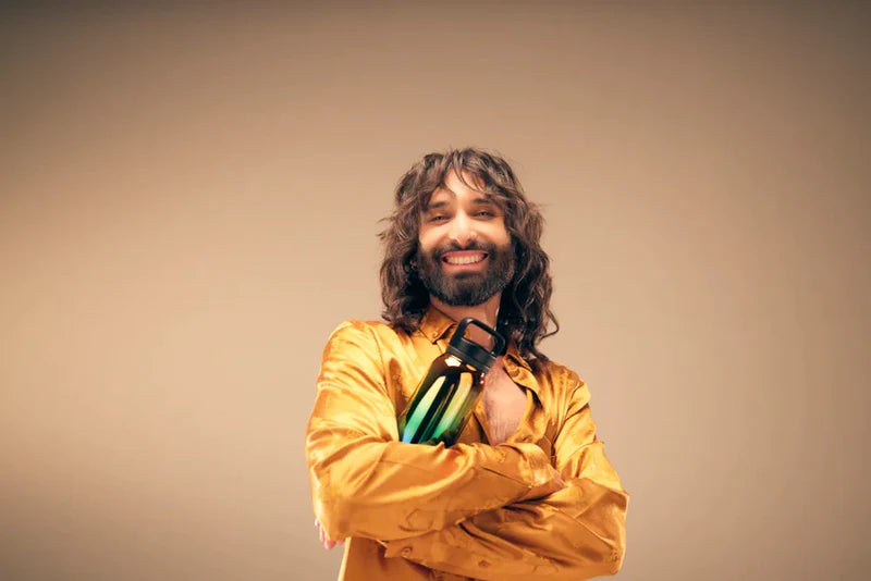A man with long hair and a beard holds a waterdrop® branded bottle indoors.