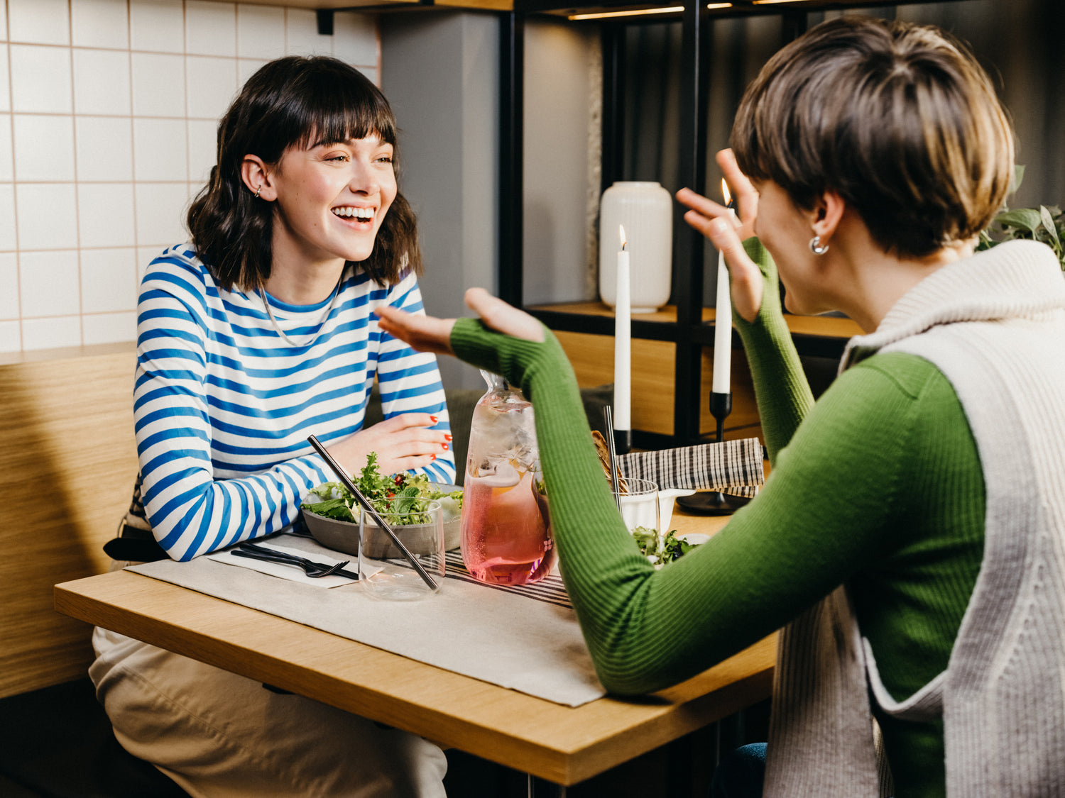 Two women sit at a table with a bowl of salad, a glass of wine, and candles, sharing a joyful moment indoors.
