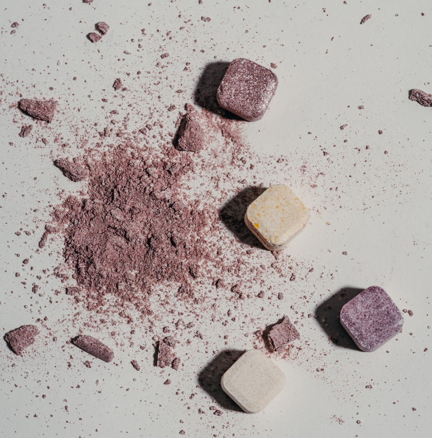 A close-up of colorful, crushed eyeshadow-like powders and small square objects, resembling waterdrop® flavored water cubes, on a white surface.