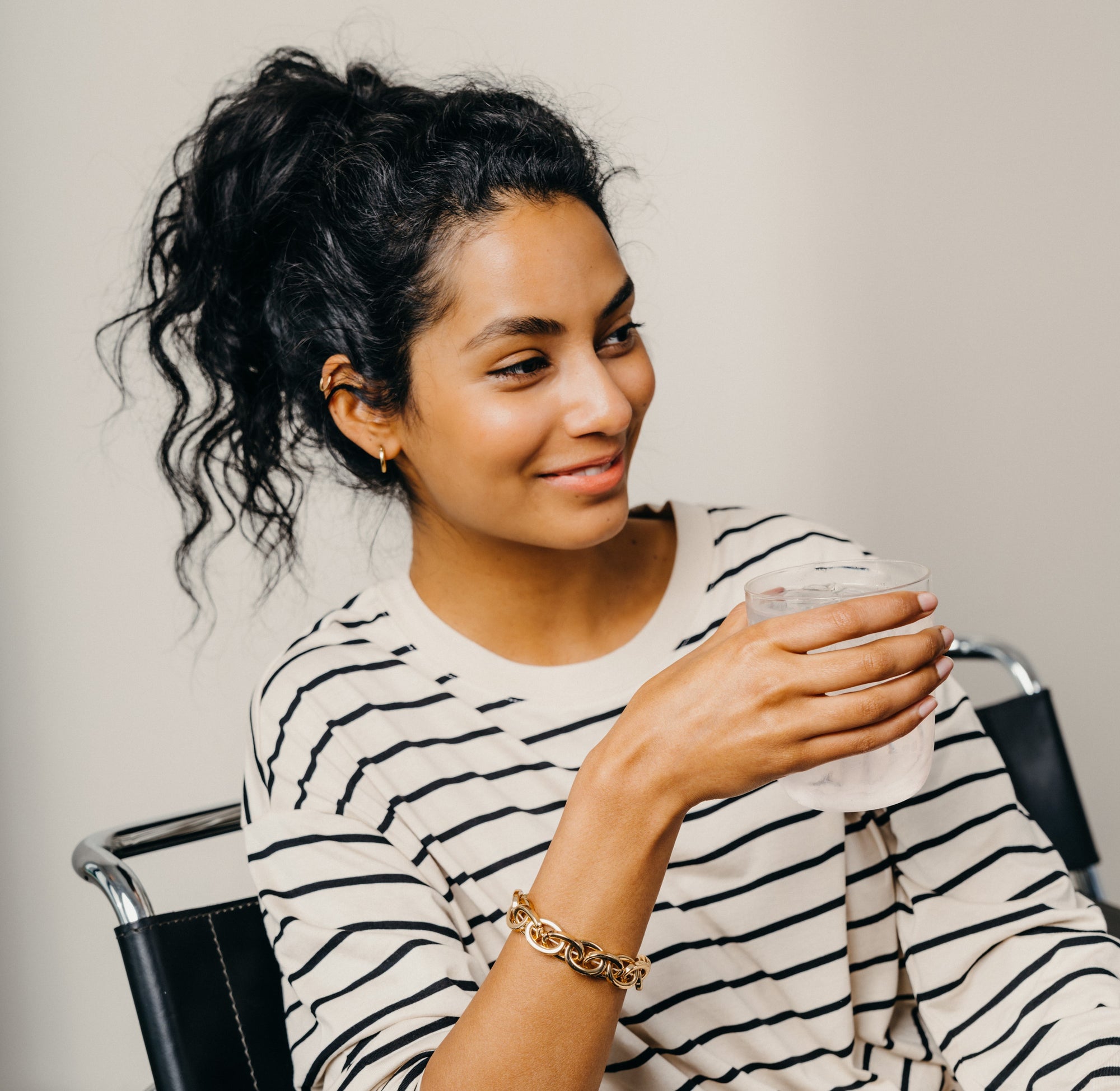 A woman with a ponytail holding a glass of water, wearing a gold chain bracelet.