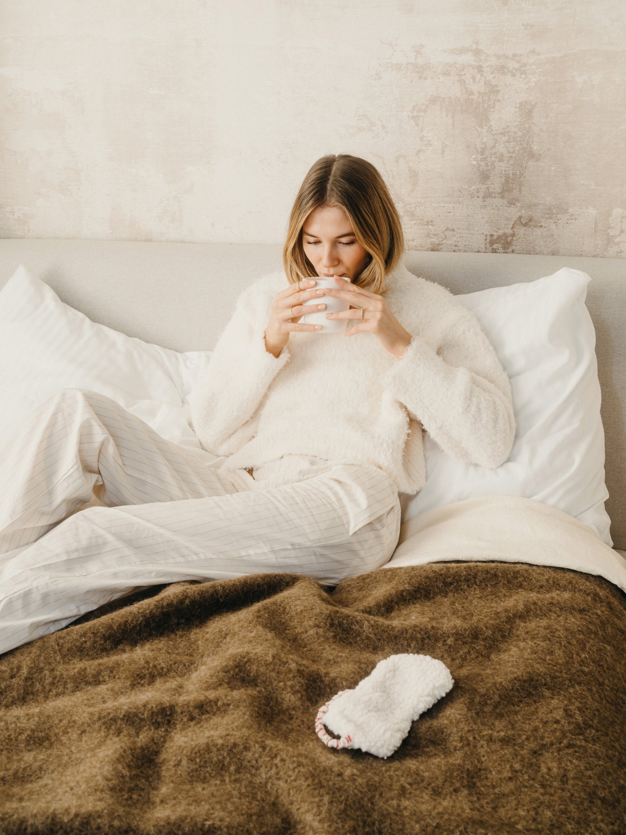 A woman sits in bed, eyes closed, holding a white cup, evoking a sense of comfort and relaxation.