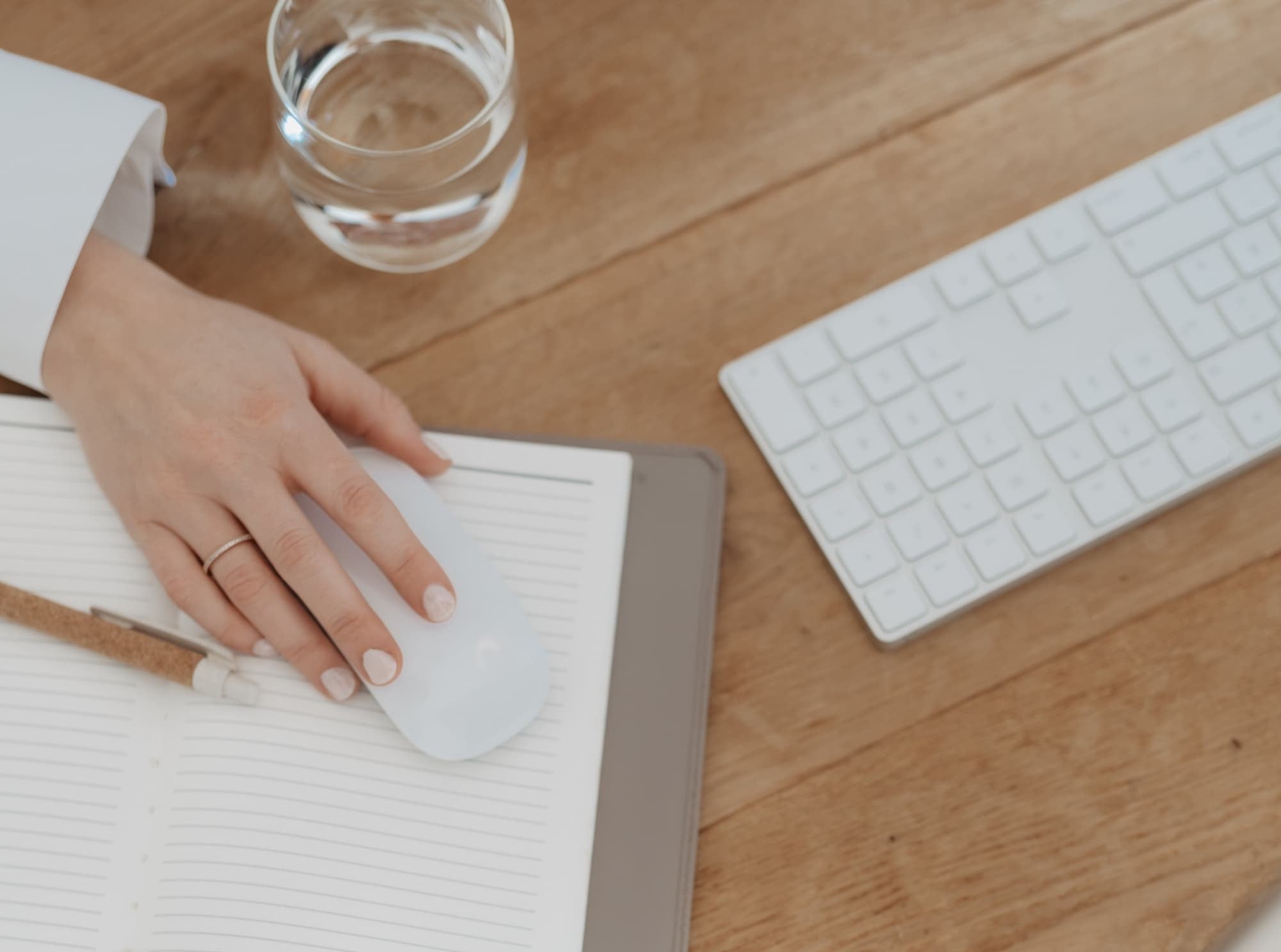 A hand using a mouse and keyboard next to a glass of water on a wooden surface.