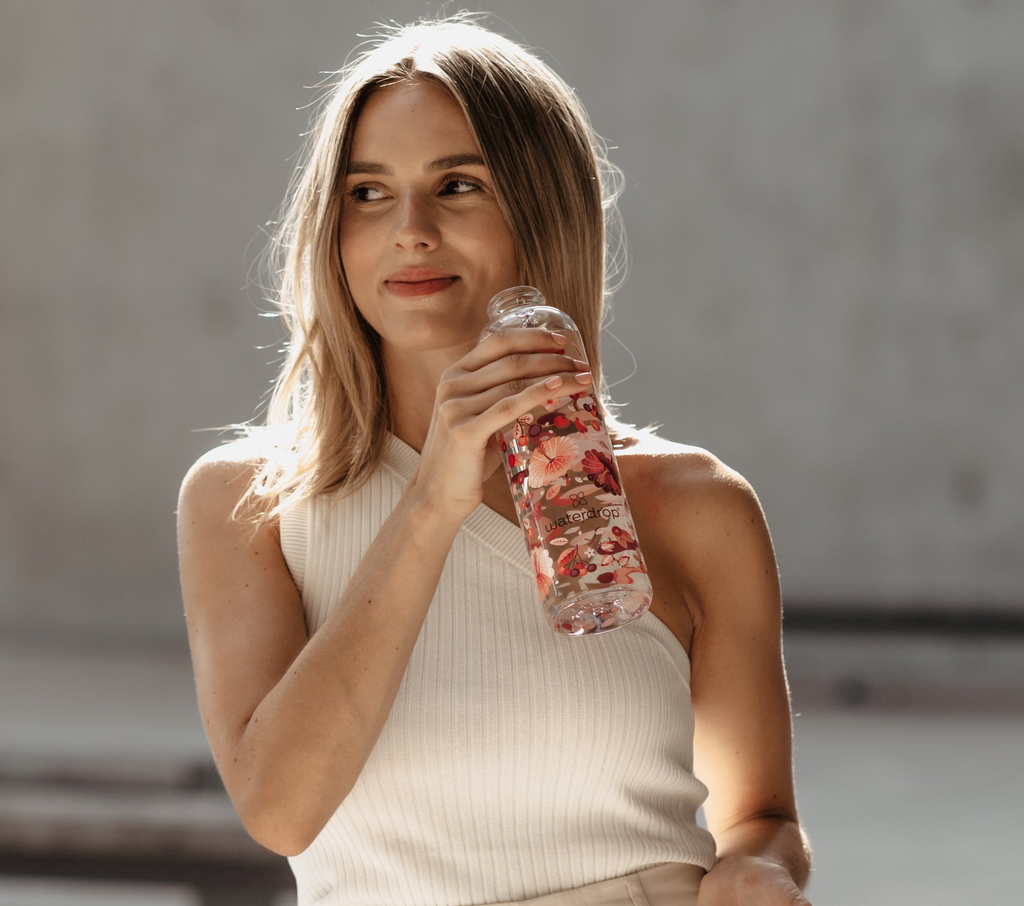 A woman holding a waterdrop® branded water bottle, wearing stylish clothing, with long hair, photographed in an outdoor setting.