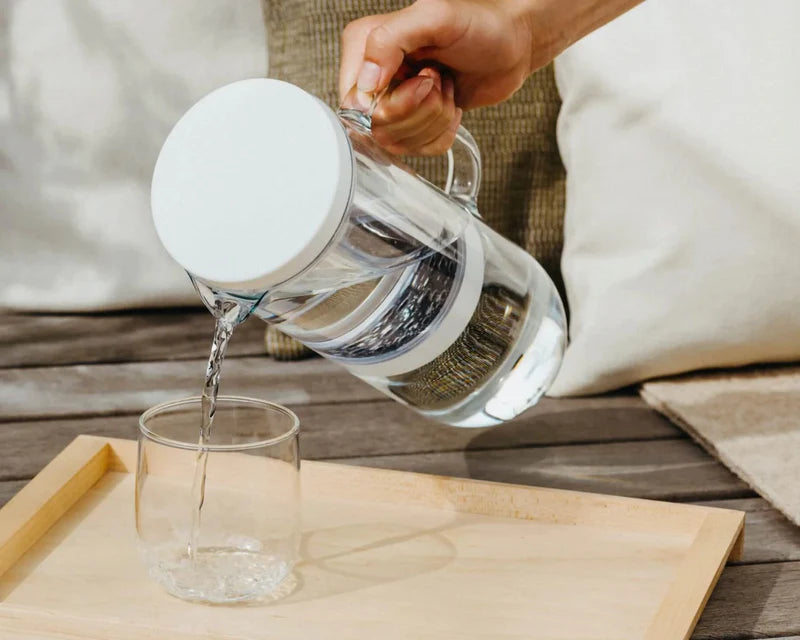 A person pouring water from a glass pitcher into a glass on a table indoors.