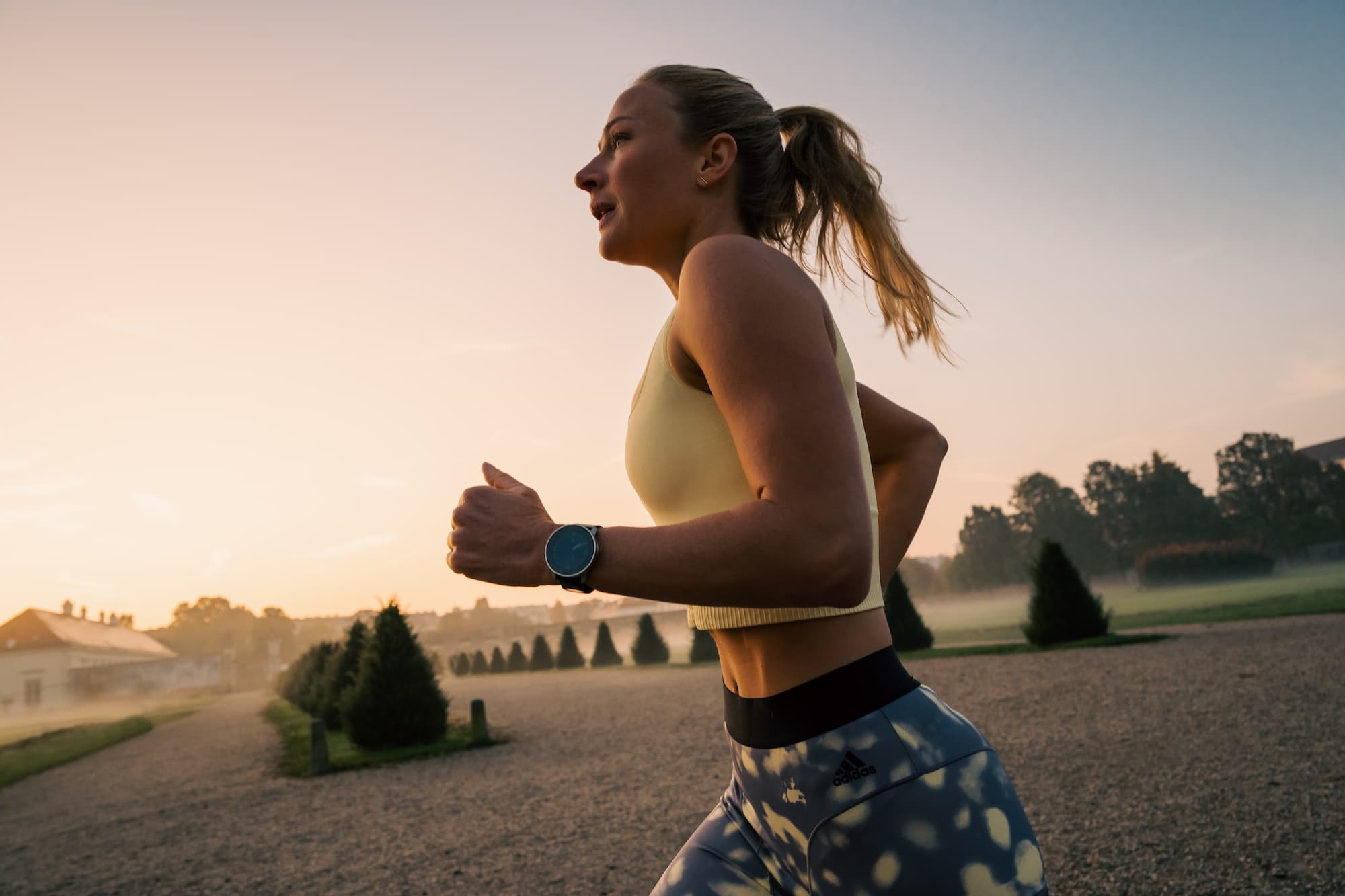 A woman running on gravel, wearing a smartwatch, in an outdoor setting with blurred trees in the background.