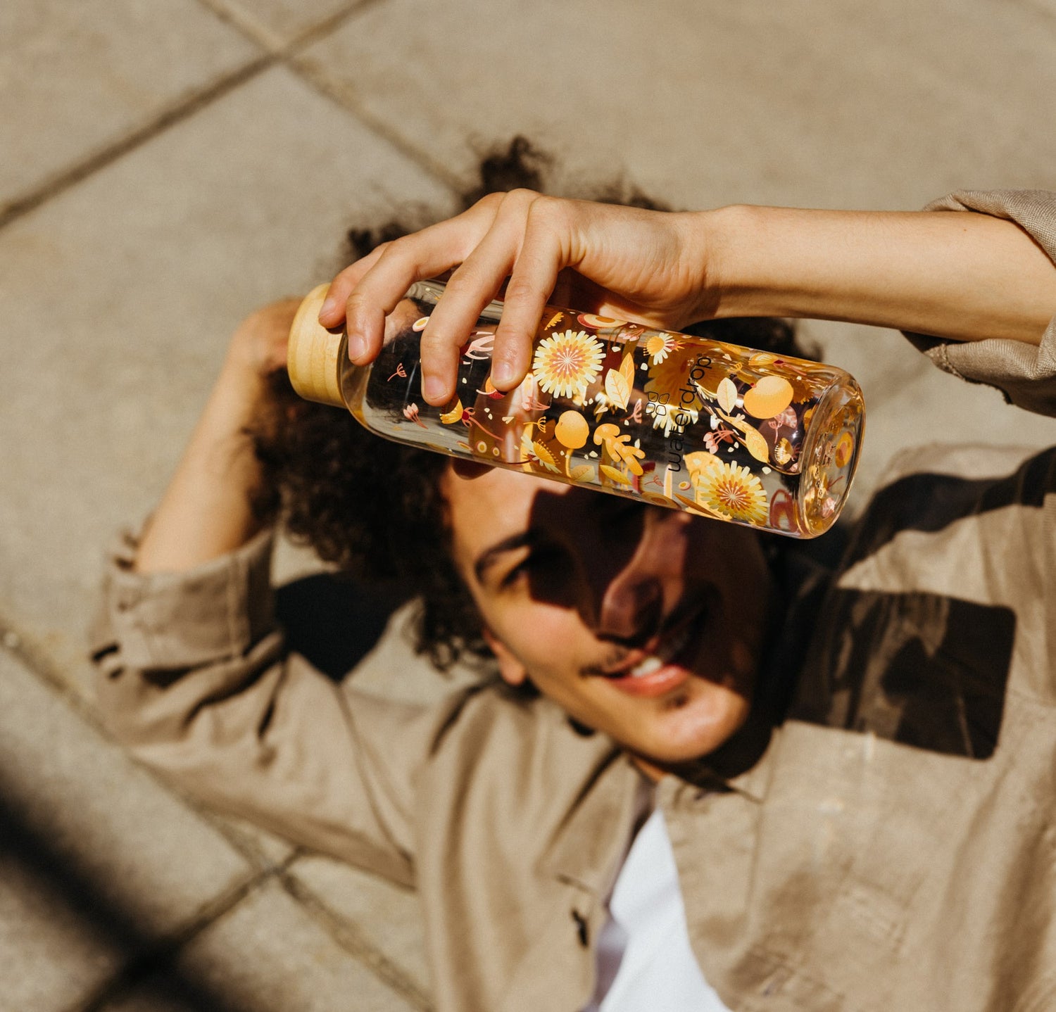 A man lying on the ground holds a waterdrop® bottle with a floral design, emphasizing the brand's focus on hydration and stylish accessories.