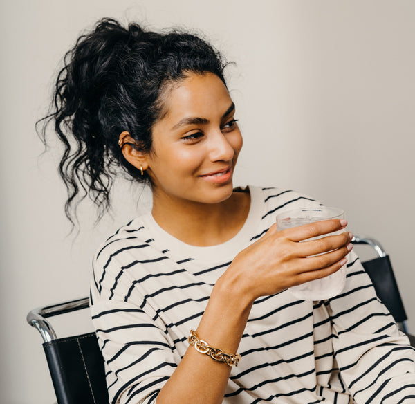 A woman with a ponytail holding a glass of water, wearing a gold chain bracelet.