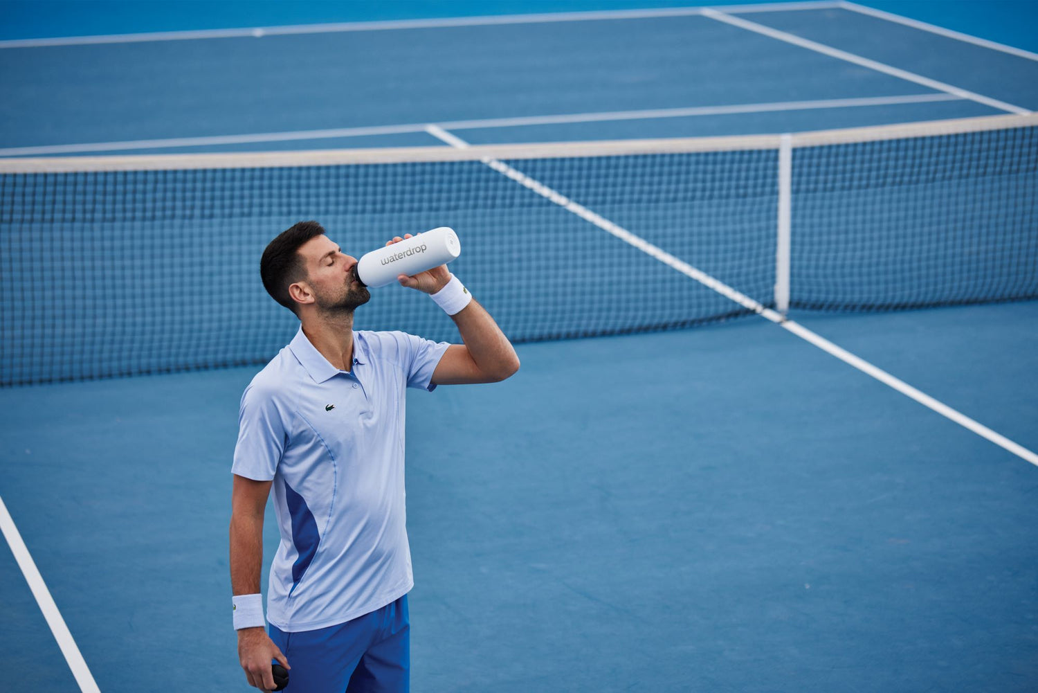 Novak Djokovic on a tennis court drinking from a white plastic cup, holding a tennis racket and wearing a white wristband.