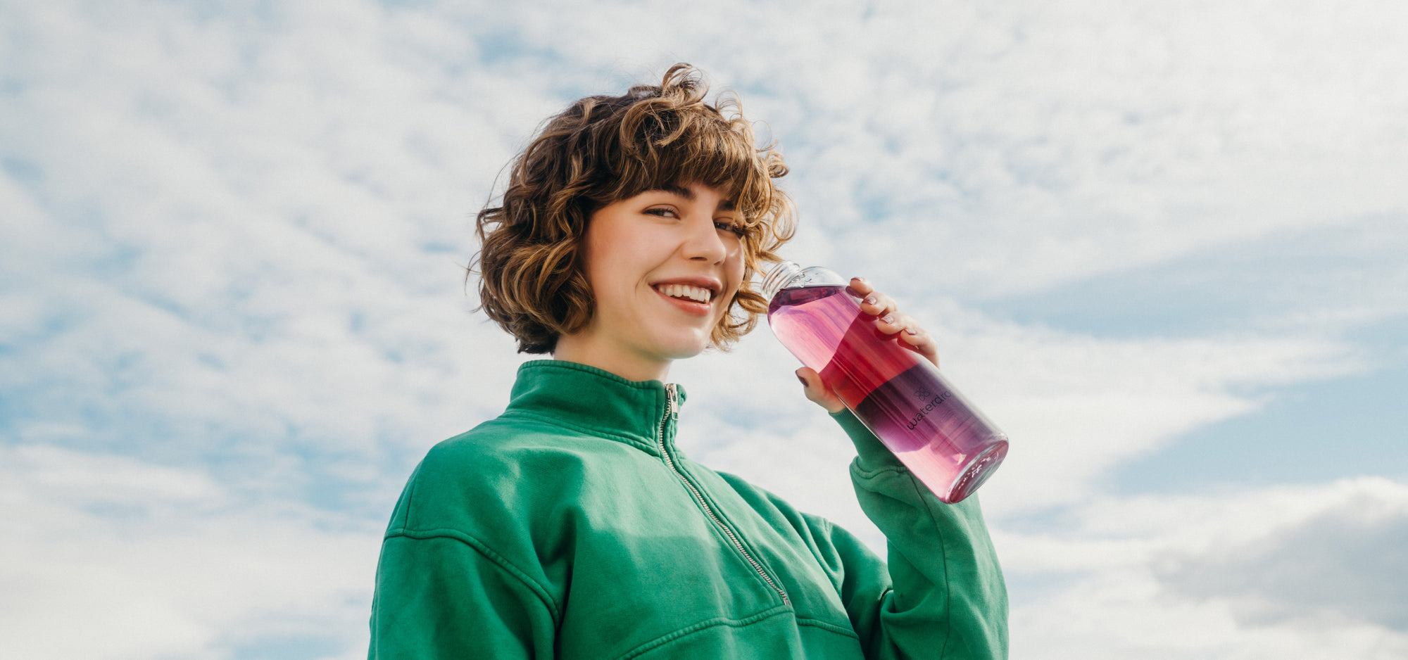 A smiling woman with curly hair holds a waterdrop® branded bottle of water.