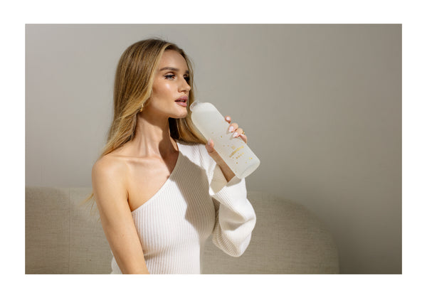 A woman with blonde hair wearing a white shirt drinks from a water bottle indoors.