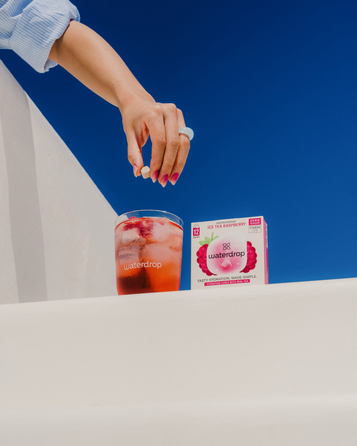 Hand dropping a hydration cube into a glass of ICE TEA from the waterdrop® ICE TEA Set, featuring LEMON, PEACH, BLUEBERRY, and RASPBERRY flavors.