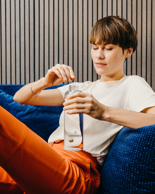 Woman sitting on a couch, holding a Microdrink Taster Pack bottle, showcasing the refreshing hydration solution with natural fruit and plant extracts.
