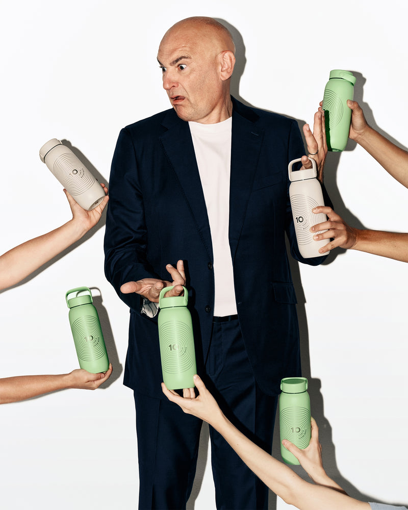 Man in a dark suit stands while several hands offer assorted green and white water bottles around him.