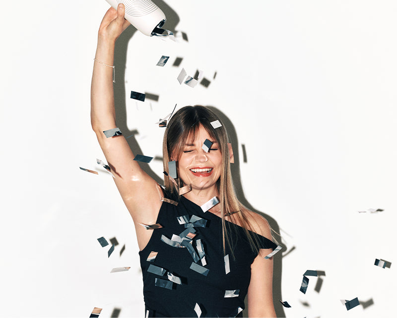 Smiling woman in a black off-shoulder dress throws confetti from a party popper.