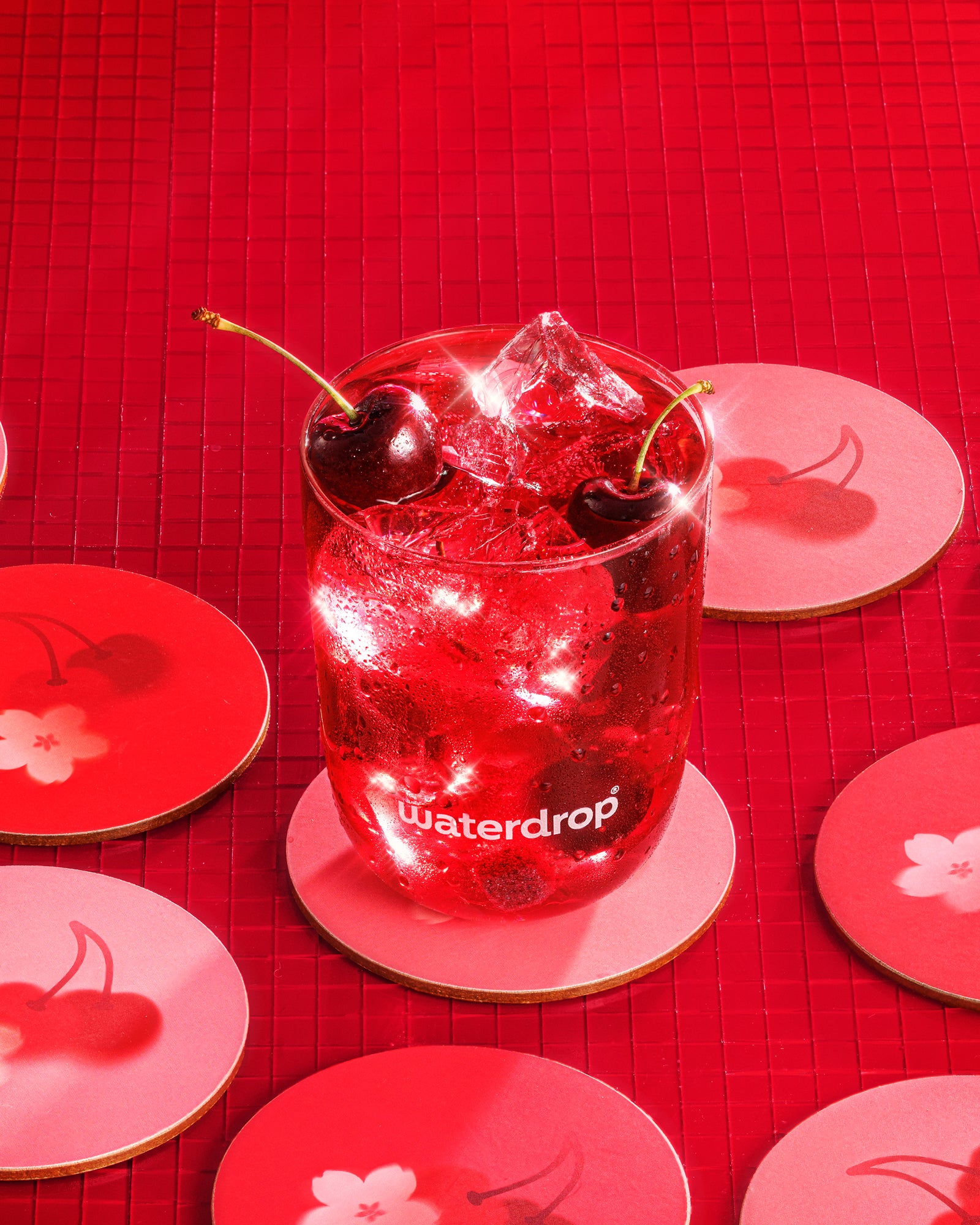 Cold red beverage with ice and two cherries in a glass on a red background with cherry-pattern coasters.