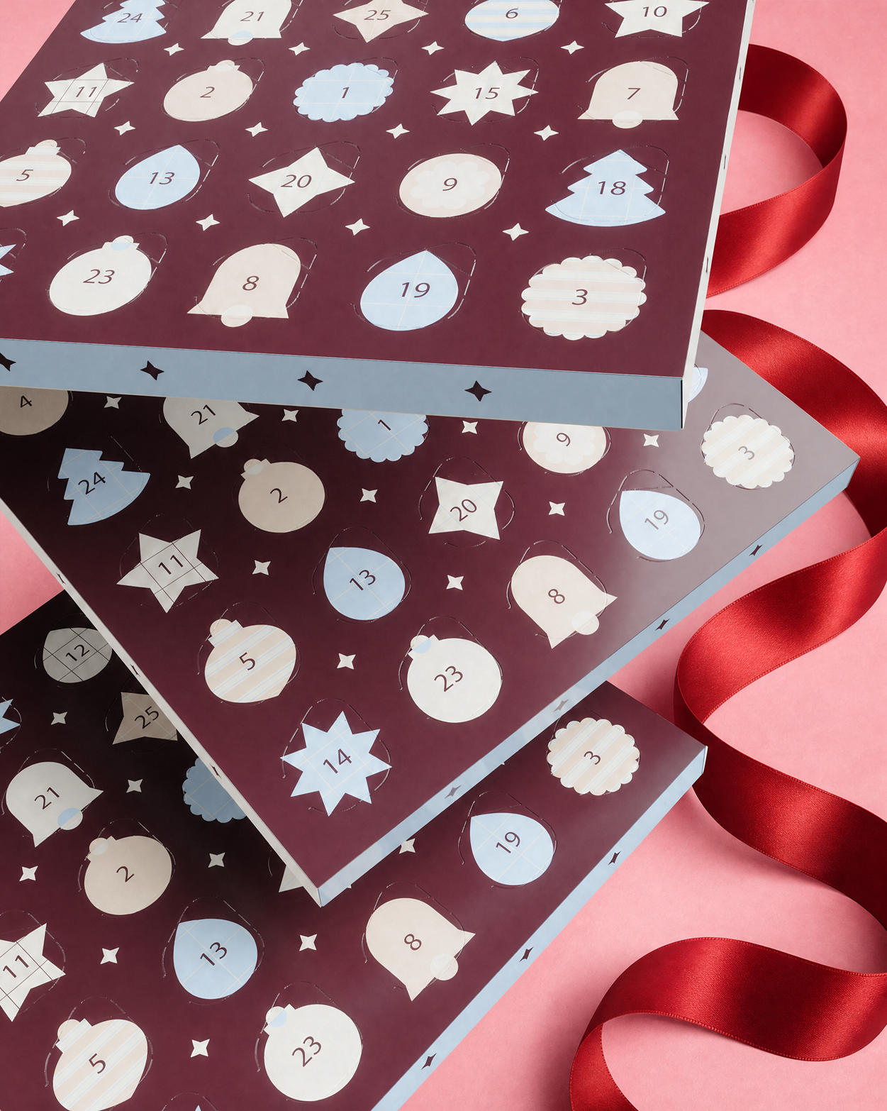 Three stacked Advent calendar boxes with numbered doors and decorative shapes, tied with a red ribbon on a pink background.