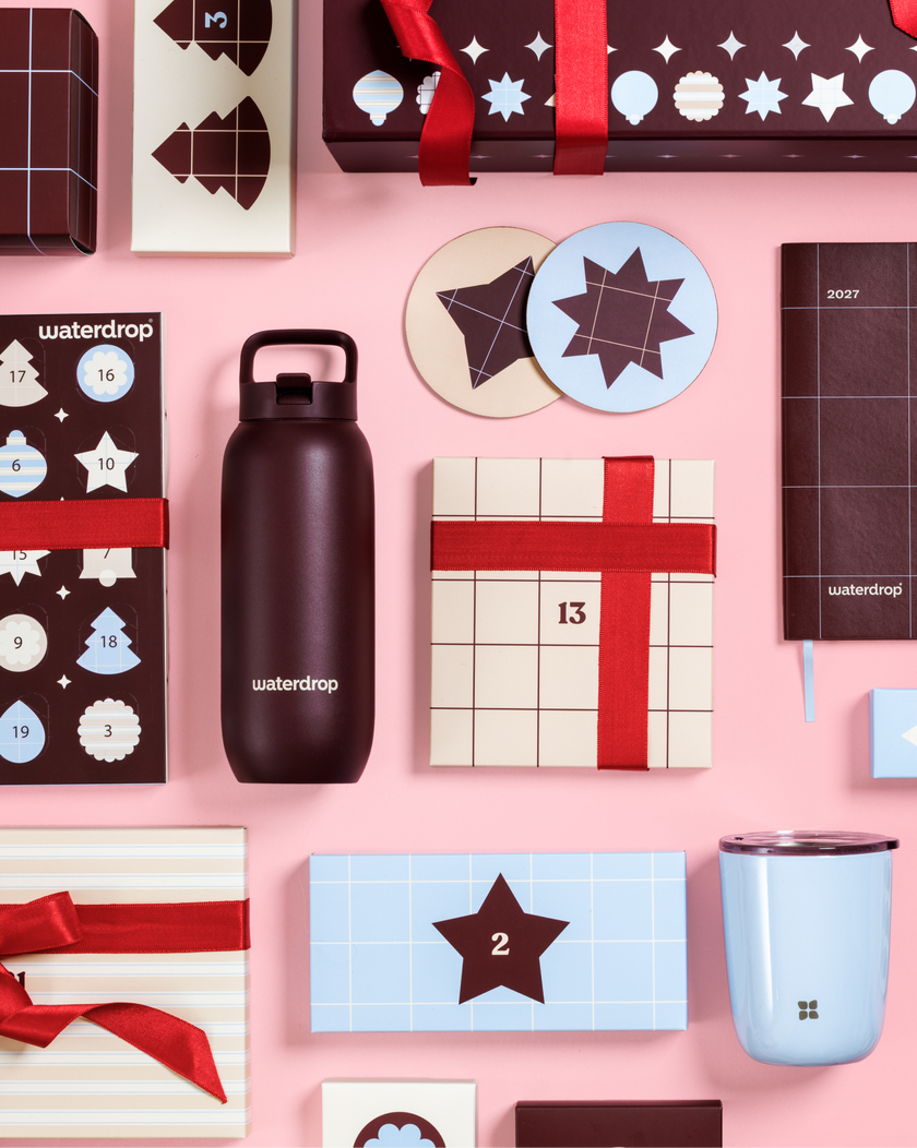 Assorted Advent calendars and gift boxes with red ribbons on a pink background, alongside a black water bottle and a light blue mug.
