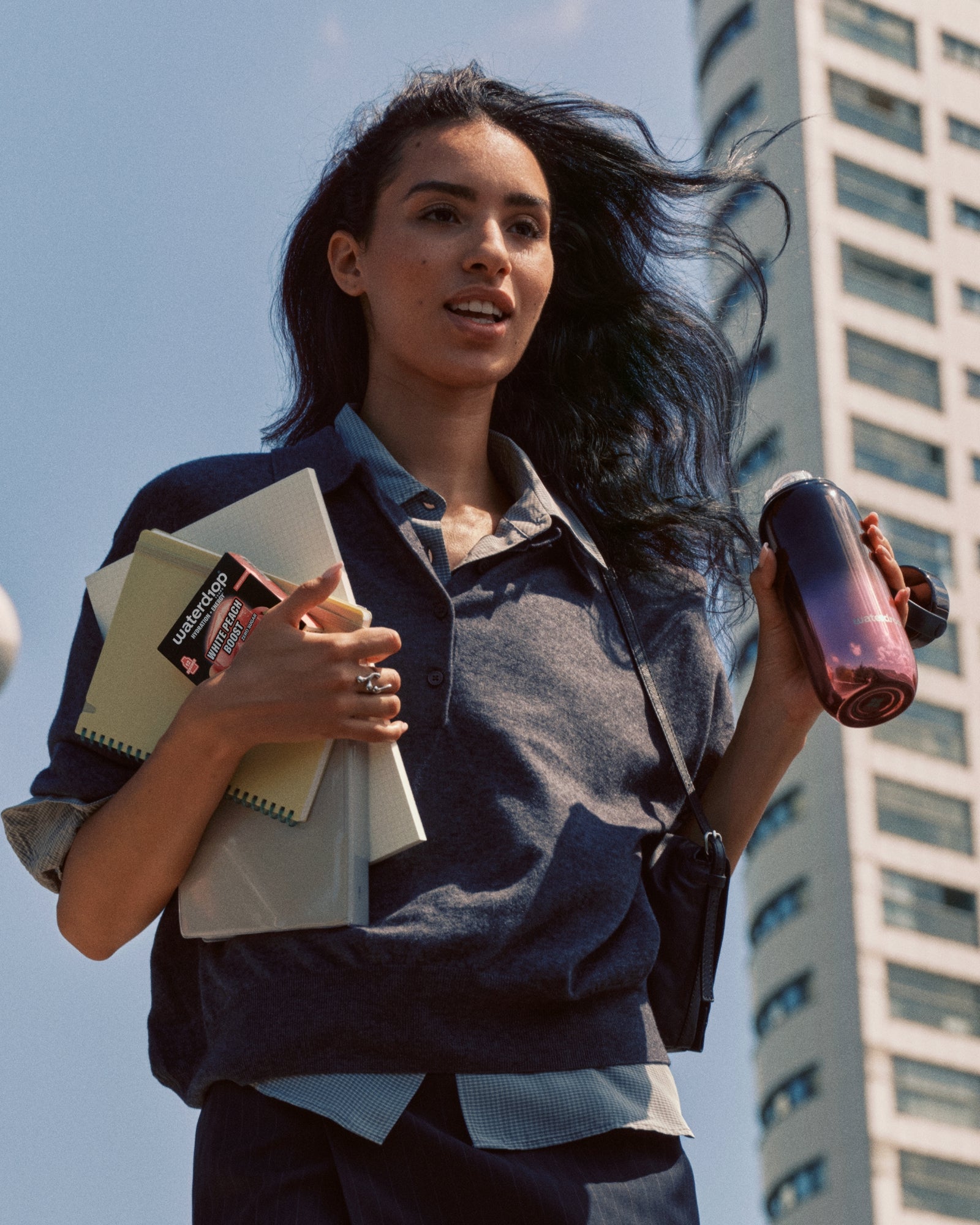 Young woman walking in an urban outdoor setting, carrying notebooks and a binder, and holding a water bottle.