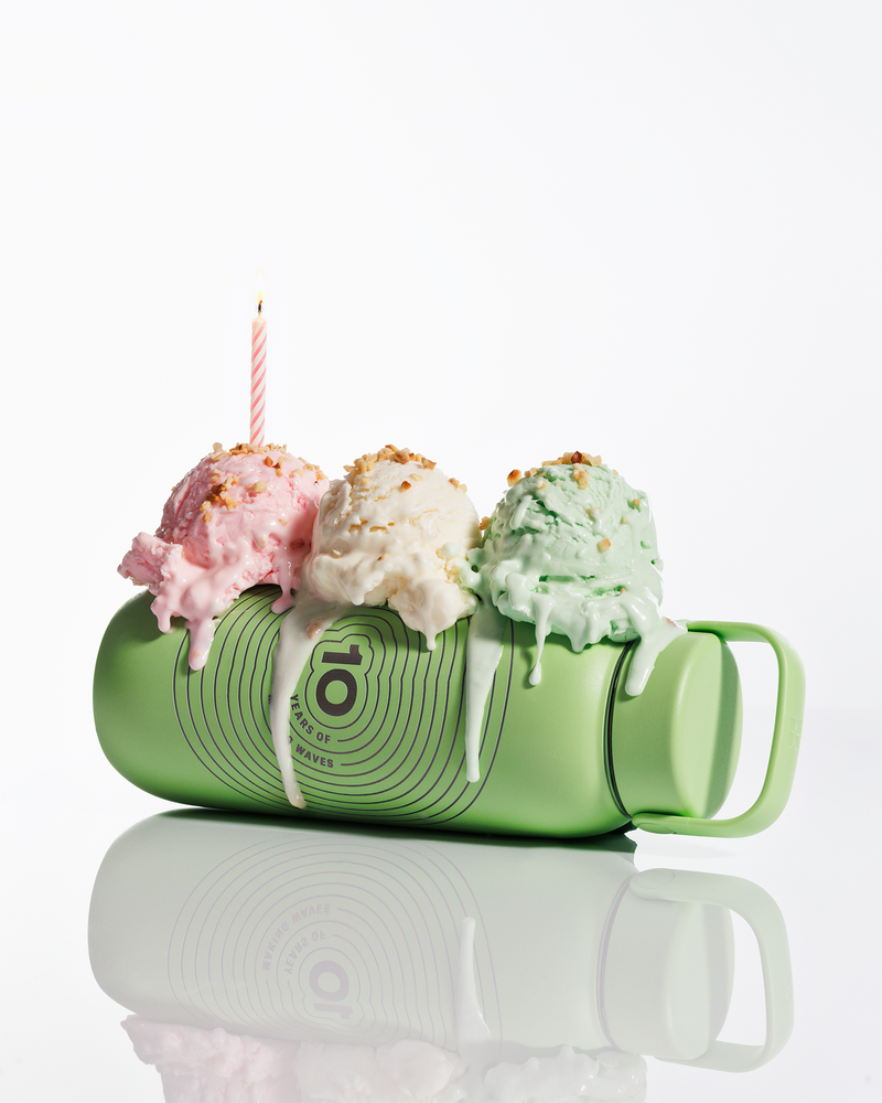 Green mug on its side with three scoops of ice cream on top and a lit candle, with melted icing dripping down.