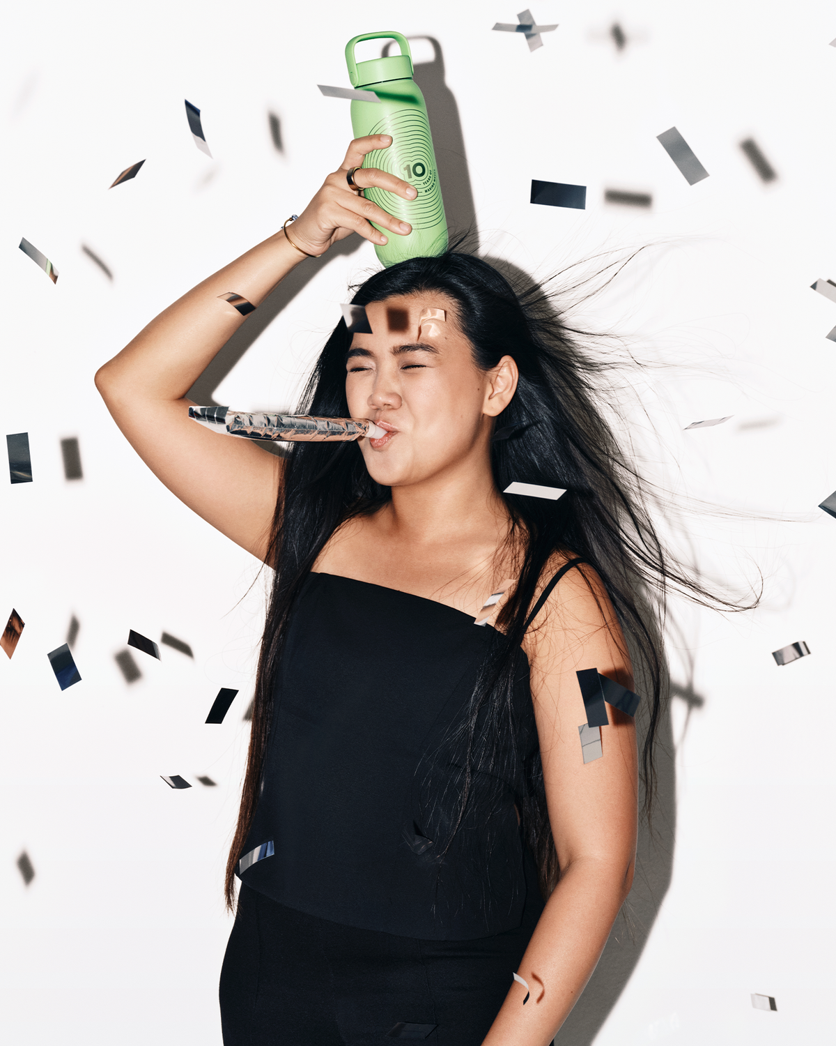 Woman in a black top holds a green spray bottle to her head as confetti falls around her.