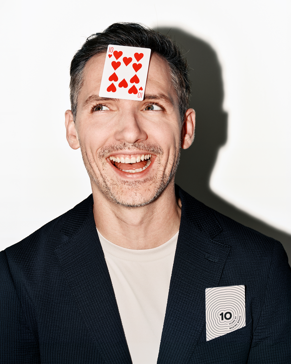 Smiling man in a dark blazer and light shirt has a playing card on his forehead and another card in his chest pocket.