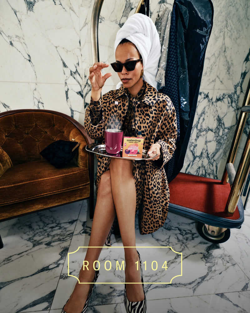 Woman in a leopard-print robe with a towel on her head sits on a marble floor, holding a tray of cosmetics.