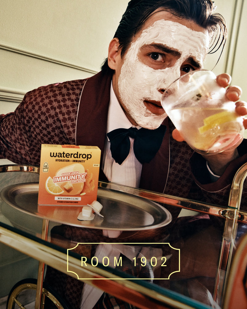 Person in a brown checked suit with a dark bow tie wearing a white face mask holds a glass of beverage beside a tray with a boxed product.