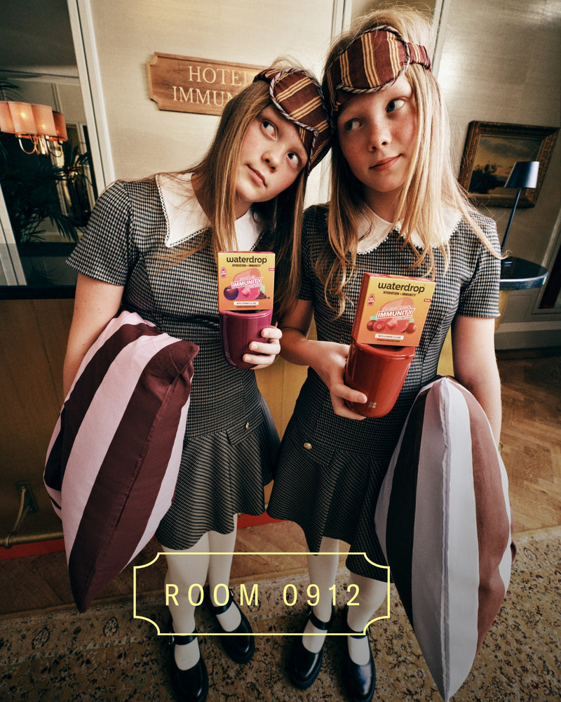 Two girls in matching outfits with plaid headbands hold colorful drinks in a hotel lobby, standing side by side with striped bags.
