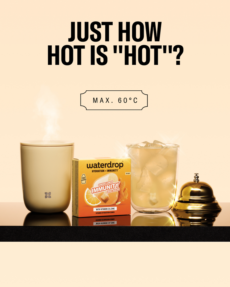 Steaming beige mug on the left, a box of Waterdrop Immunity lemon drink, a glass with ice, and a small gold bell on a black shelf.