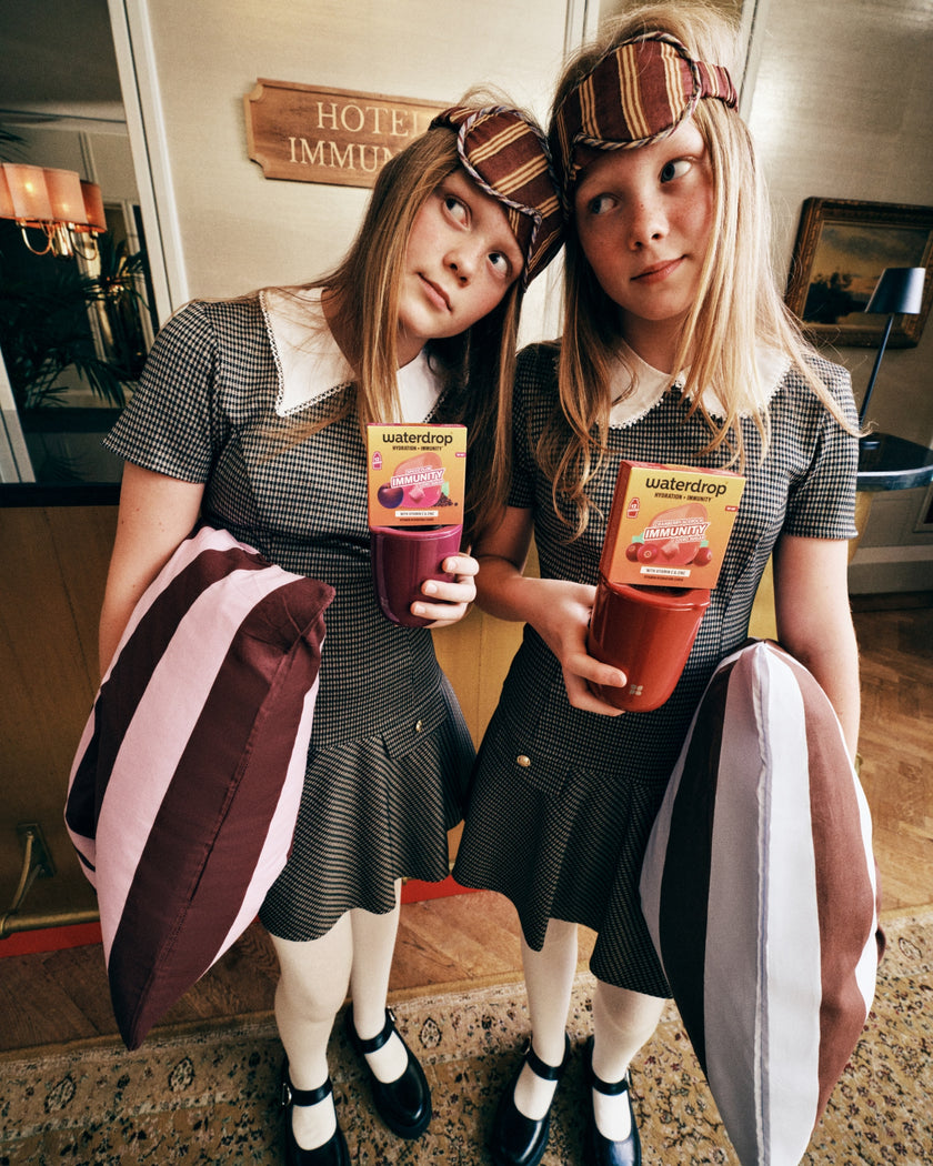 Two girls in matching grey dresses with white collars hold juice drinks in a hotel lobby.