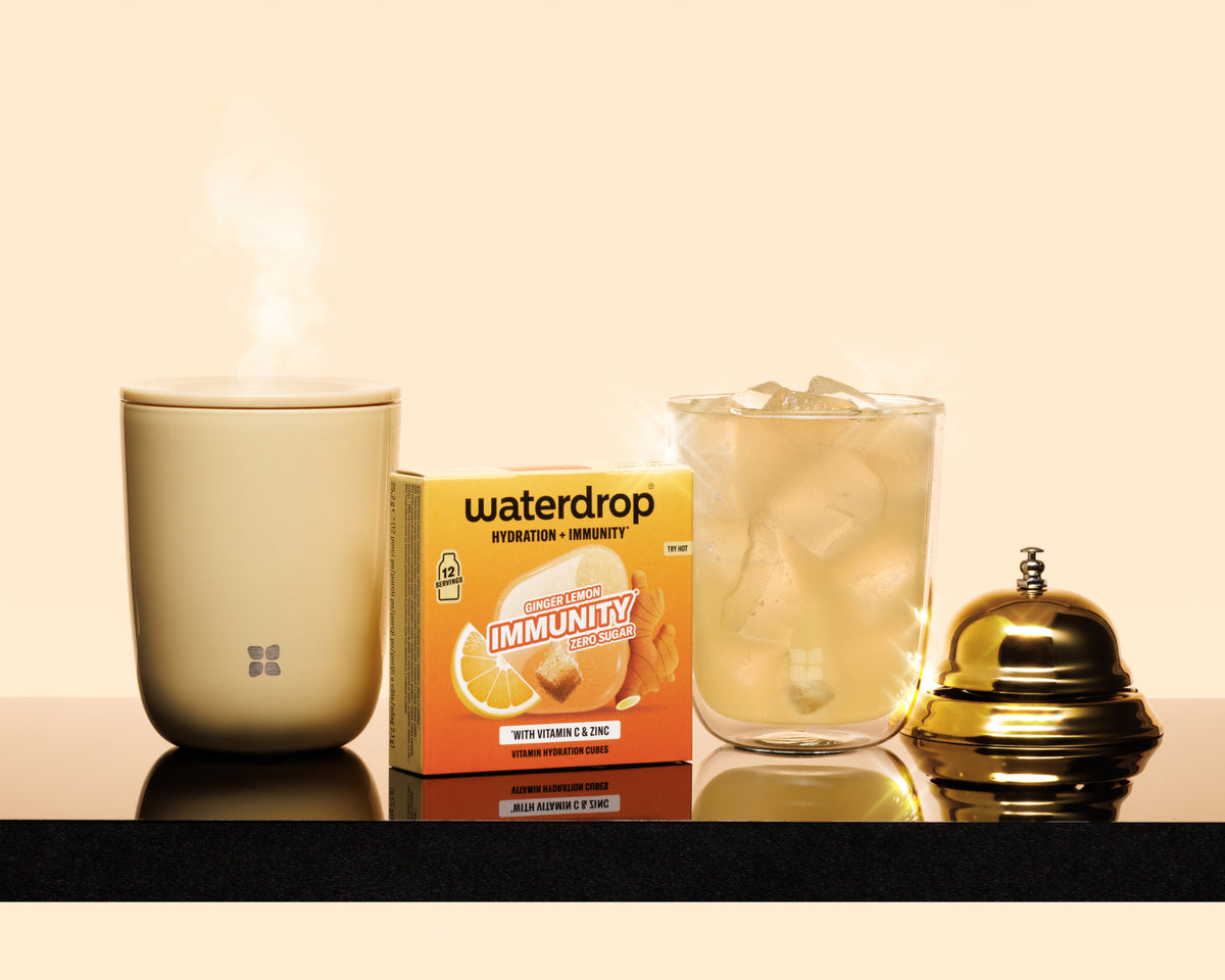 Steaming beige mug, boxed immunity product, glass with ice, and a polished gold bell on a black counter.
