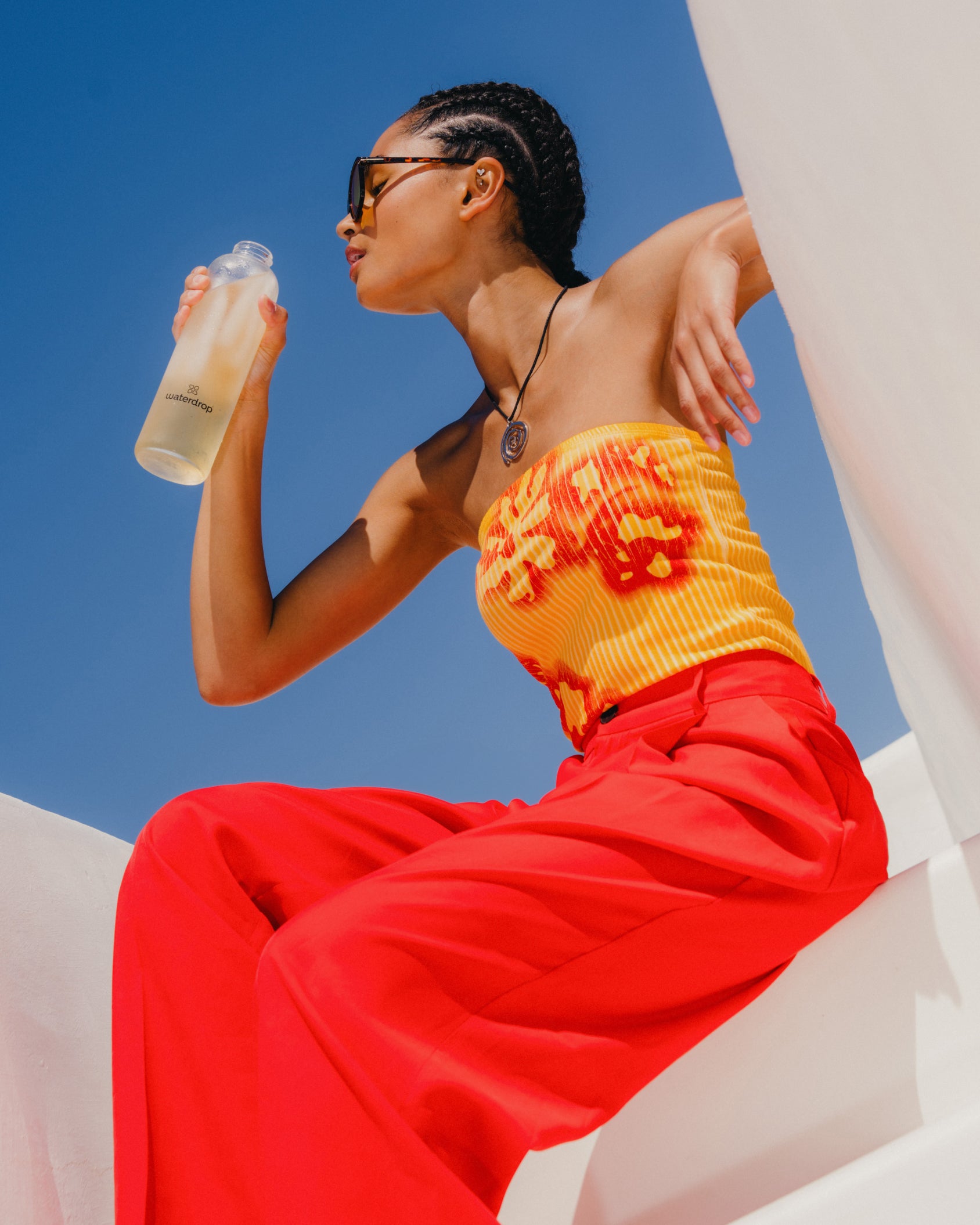 Woman sitting on a ledge, drinking ICE TEA Peach from a waterdrop® bottle, enjoying a refreshing black tea with a fruity twist, sugar-free.