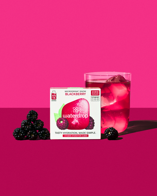 Glass of iced pink BLACKBERRY Microdrink next to waterdrop 12-pack, accompanied by fresh blackberries.	