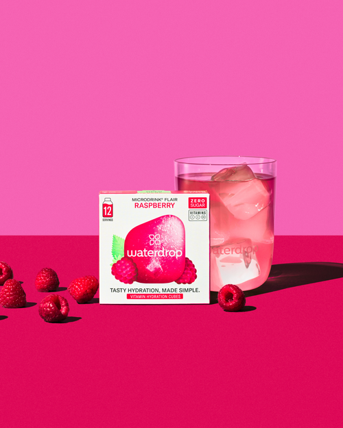 Box of RASPBERRY flavored water cubes beside a glass of pink drink with raspberries and ice, highlighting the refreshing blend of raspberry, elderflower, and lime blossom.