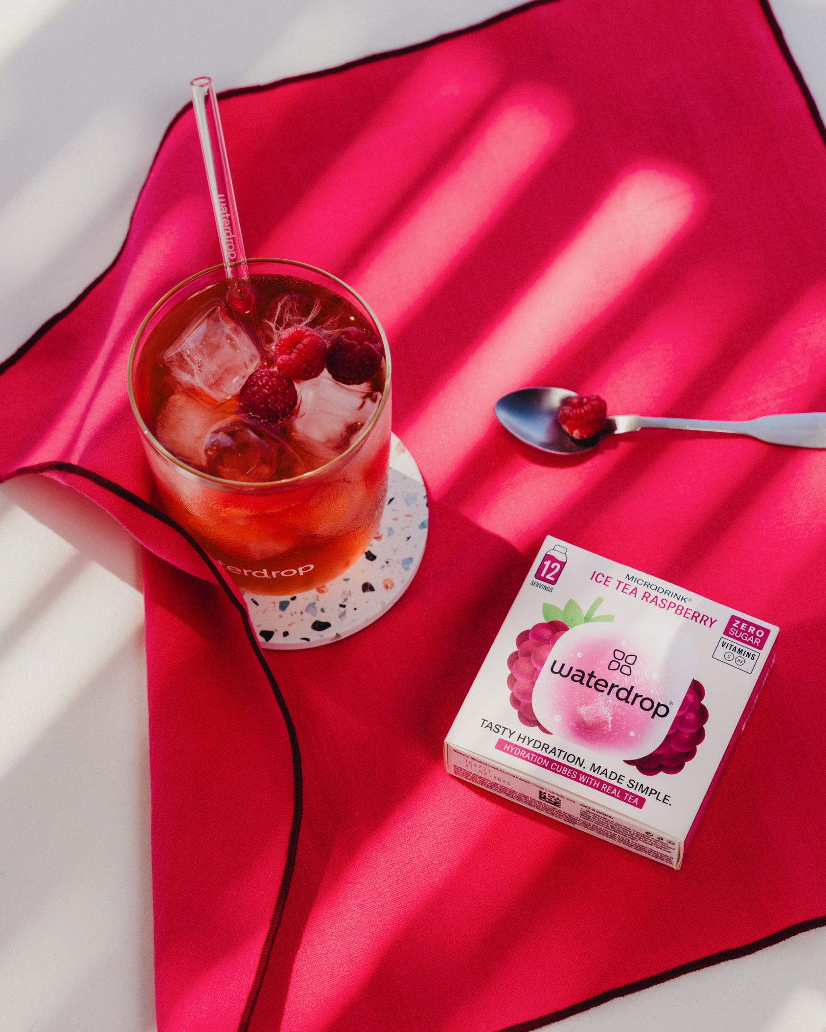 Glass of ICE TEA RASPBERRY with ice cubes and raspberries, placed next to a box of the product on a red surface.